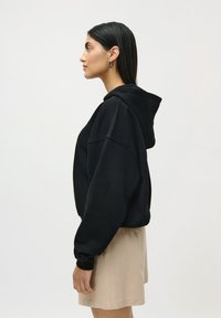 Woman with straight black hair wearing silver earrings, black hoodie, and beige skirt standing in profile against a plain background.
