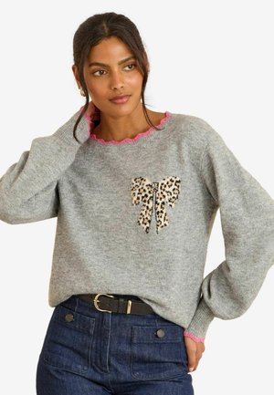 Grauer Pullover mit langen Ärmeln, pinkem gewelltem Ausschnitt und Leo-Print-Bow-Applikation auf der Brust. Kombiniert mit dunkelblauen Jeans und einem Gürtel.