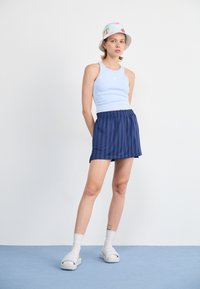 Lyseblå tanktop med logo, marineblå stripete plisserte miniskjørt, hvite sandaler med åpen tå, og mønstret bøttehatt. Glatt stofftekstur.