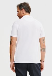 Biała koszulka polo z krótkim rękawem, klasycznym kołnierzykiem i rozcięciami po bokach u dołu, prezentowana na modelu widokiem z tyłu.