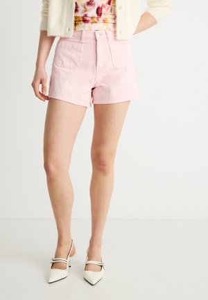 Donna che indossa pantaloncini rosa chiaro a vita alta, una maglietta motivo fantasia, un cardigan color crema e décolleté bianchi con punta a punta e cinturini alla caviglia.