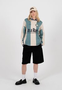 Persoană purtând un pulover în dungi albastre și bej cu text, pantaloni scurți negri, șosete albe, pantofi loafer negri și o șapcă bej cu text, stând în fața unui fundal alb.