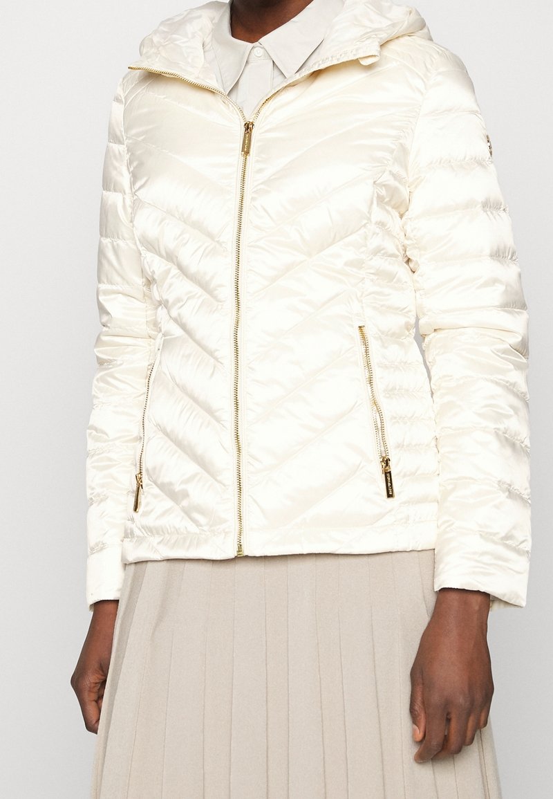 Puchowa kurtka w kolorze kości słoniowej z kapturem, z charakterystycznym wzorem w zygzak, złotym zamkiem błyskawicznym i bocznymi kieszeniami. Gładka faktura i lekki materiał.