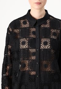 Svart spetsblus med rutmönster och blomsteraccenter, knappstängning fram och klassisk kragsdesign.