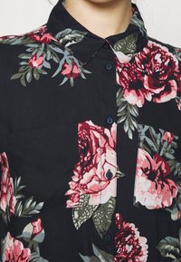 Camicia floreale nera con motivi di fiori rosa e verdi. Presenta un colletto, chiusura con bottoni e taschino sul petto. Tessuto morbido.