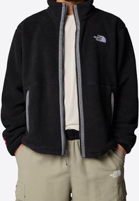 Svart fleecejacka med hög krage, dragkedjestängning och grå dekorativ sömnad. Har sidofickor och logotyp på övre vänstra bröstet.