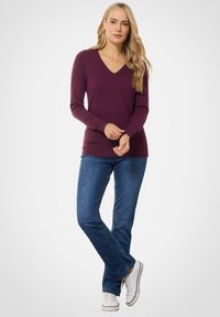 Pruimen V-hals trui met lange mouwen, gecombineerd met blauwe flared jeans en witte sneakers. Soepele stof, getailleerde snit en ribbelzoom.