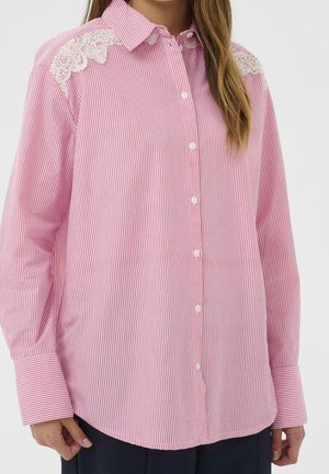 Femme portant une chemise à fines rayures roses et blanches à manches longues avec des accents en dentelle blanche aux épaules, boutonnée sur le devant.