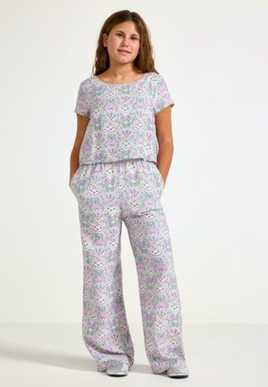 Tyttö seisoo käsineen taskuissa pukeutuneena laventelin, vihreän ja valkoisen kuvioituun lyhythihaiseen yläosaan ja leveälahkeisiin housuihin hopeisten kenkien kanssa.