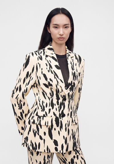 Femme aux longs cheveux noirs porte un ensemble blazer et pantalon beige avec de grandes taches noires abstraites sur un haut noir, debout devant un fond uni.