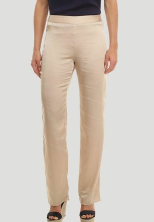Mujer con pantalones beige de corte recto y sandalias negras de cuña con los dedos abiertos, de pie frente a un fondo liso y claro.