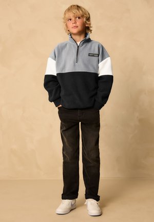 Blond jongetje draagt een grijze, witte en zwarte fleece met rits, zwarte spijkerbroek en witte sneakers, staat met handen in de zakken.