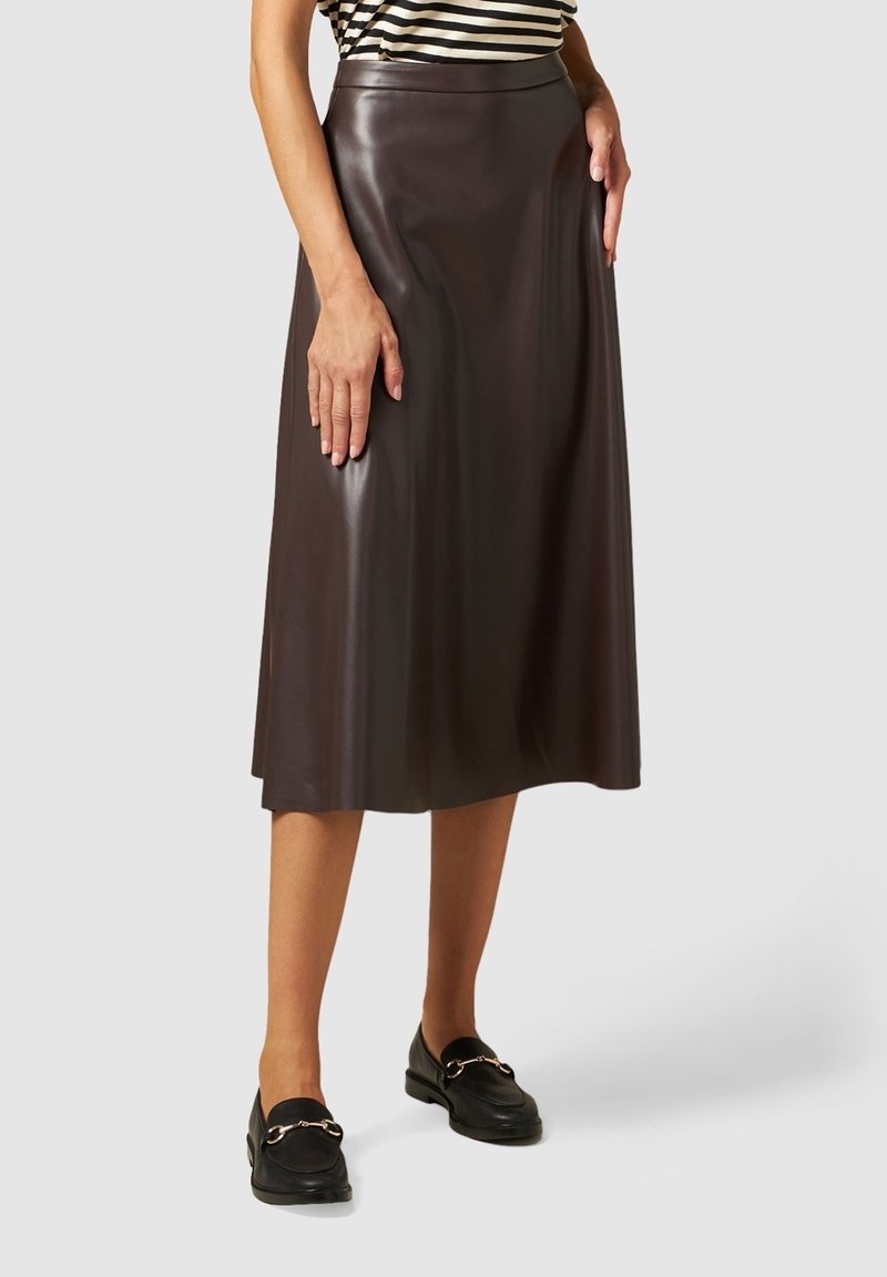Femme portant une jupe midi en cuir marron foncé taille haute, des mocassins noirs et un haut rayé, debout devant un fond uni.