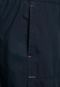 Pantaloni corti navy realizzati in tessuto leggero, caratterizzati da cuciture rosse a contrasto e un piccolo logo metallico. Texture liscia con dettagli di tasca laterale.
