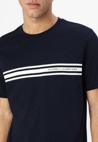 Tumesinane puuvillane T-särk, mis on kaunistatud horisontaalse valge triibuga, mille keskel on tekst "HILFIGER Est. 1985 UUS YORK". Ümar kaelus.