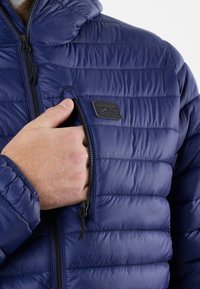Marineblauwe pufferjas met een capuchon, voorzien van een horizontaal gestikte ontwerppatroon, een ritszak aan de voorkant en een klein zwart logo-patch op de borst.