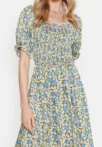 Kukallinen mekko, jossa on röyhelörintaosa, puffihihat ja täysimittainen hame. Väreinä sinisiä, keltaisia ja valkoisia kukkia tummalla taustalla.