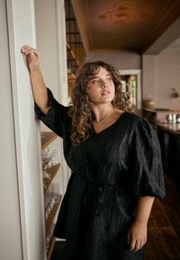 Musta, teksturoitu mekko, jossa on V-pääntie ja puffihihat, vyötäröltä kiristettävä solmulla. Pehmeä, laskeutuva kangas, jossa on hienovarainen kiilto.