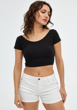 Mujer con cabello rizado que lleva una camiseta negra corta y pantalones cortos blancos, posando contra un fondo claro y sencillo.