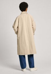 Beige dlouhý kabát s uvolněným střihem, vyrobený z lehkého materiálu. Obsahuje klasický límec a detail zpětného rozparku, kombinovaný s modrými džíny.