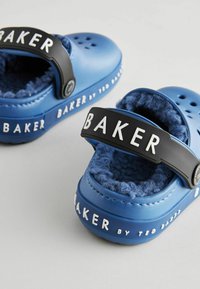 Plave gumene klompe s crnim trakama koje sadrže bijeli tekst "BAKER". Unutra meka, teksturirana plava podstava. Dizajn s više rupa za prozračnost.