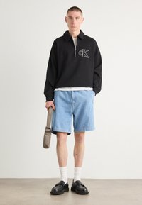 Pullover nero con zip e colletto, logo bianco, abbinato a pantaloni shorts in denim azzurro chiaro, calze bianche e mocassini neri, mentre tiene una borsa grigia.