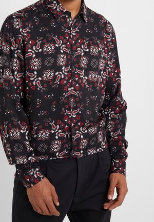 Mann trägt ein schwarzes Hemd mit roten und weißen komplexen Blumen- und Paisleymustern, kombiniert mit schwarzen Hosen.