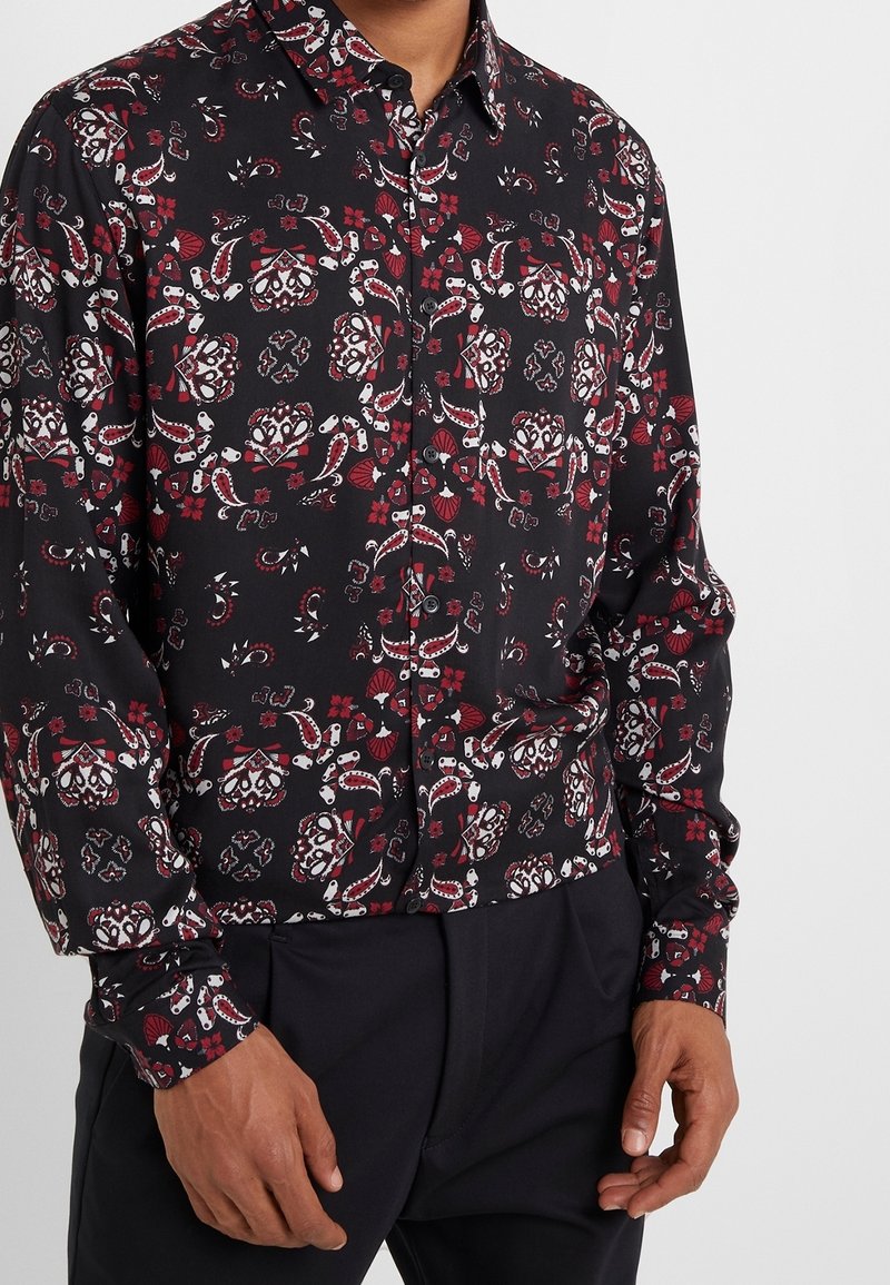 Homme portant une chemise noire à boutons avec des motifs floraux et cachemire rouges et blancs complexes, associée à un pantalon noir.