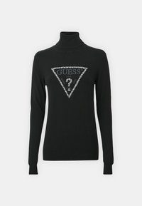 Maglione nero a collo alto con design aderente, caratterizzato da un logo triangolare argentato scintillante con la scritta "GUESS" al centro. Maniche lunghe.