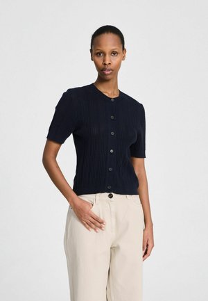 Žena s krátkymi tmavými vlasmi, oblečená v čiernej krátkosvetej pletenine na gombíky a béžových vysokých nohaviciach, stojaca s jednou rukou v kapse.