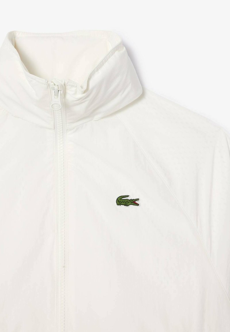 Biała kurtka zapinana na zamek błyskawiczny z wysokim kołnierzem i małym, zielonym logo krokodyla wyhaftowanym po lewej stronie klatki piersiowej.