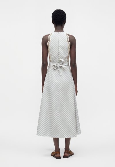 Femme portant une robe midi rayée sans manches avec un bord en dentelle et un nœud noué à la taille, associée à des sandales plates marron, debout de dos.