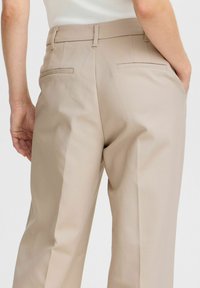 Pantalons beige sur mesure avec une texture lisse, dotés de poches arrière et d'une coupe straight leg. Détails de couture subtils sur la taille.