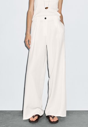 Woman wearing white wide-leg pants, white sleeveless top, brown flip-flops, and a necklace with a brown pendant, standing on gray floor.