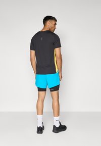 Sort og gul kortærmet træningsskjorte med rund halsudskæring, parret med turkise shorts. Outfitet inkluderer sorte sneakers og hvide sokker.