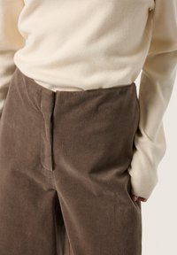 Brown corduroy pants with a high waist and vertical ribbed texture, paired with a cream-colored long-sleeve top, close-up view.