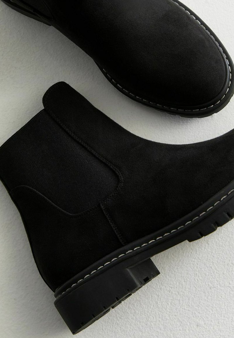 Schwarze Wildleder-Stiefeletten mit elastischen Seitenpaneelen, runder Zehenpartie und strukturierten Gummisohlen. Kontrastierender weißer Näht entlang der Kante.