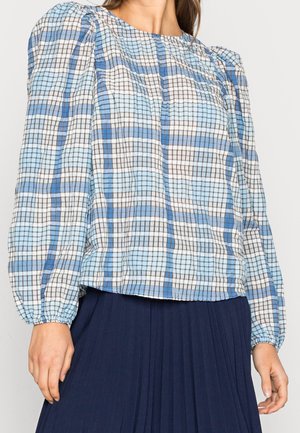 Femme portant un chemisier à carreaux bleu et blanc avec des manches longues bouffantes et une jupe plissée bleu foncé.