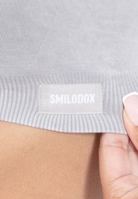 Vaalea harmaa vaatekangas, jossa on palkkirakenteinen pinta, ja valkoinen etiketti, jossa on brändin nimi "SMILODOX" ommeltuna yhteen sivuun.