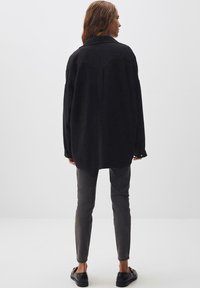 Camicia nera oversize con colletto, maniche lunghe e cucitura sul retro; abbinata a pantaloni grigi aderenti e scarpe nere.