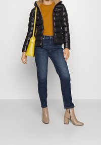 Černá péřová bunda, hořčicová košile, tmavě modré džíny, béžové kotníkové boty na blokovém podpatku a žlutá kabelka přes rameno. Hladké textury, ležérní styl.