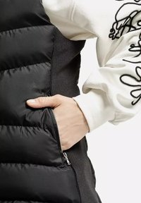 Czarna kamizelka puchowa z kieszenią na zamek, z teksturowanym panelem bocznym i białą koszulą z długim rękawem z czarnymi detalami.