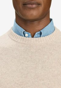 Beige gebreide trui met een geribde halslijn, gedragen over een lichtblauw hemd met knopen. Opvallende kraag en een soepele textuur.