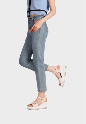 Donna che indossa jeans azzurri corti e sandali beige con plateau, in piedi con una gamba piegata su uno sfondo neutro.