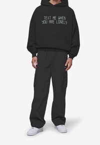 Sweat à capuche noir avec texte vert indiquant « ENVOIE-MOI UN MESSAGE QUAND TU ES SEUL. » Associé à un pantalon cargo noir ample et des baskets blanches.