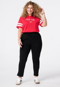 T-shirt en coton rouge avec des rayures blanches sur les manches ; présente un texte graphique. Pantalons noirs fuselés avec taille à cordon de serrage. Baskets blanches avec des rayures noires.