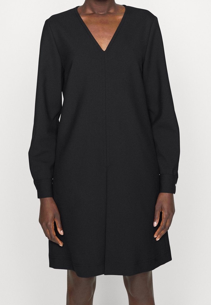 Femme portant une robe noire à manches longues, longueur genoux, avec un décolleté en V profond, debout sur un fond clair uni.