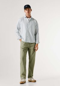 Lichtblauw geruit overhemd met lange mouwen, groene broek en beige sneakers. Zachte stof, comfortabele pasvorm en klassieke kraag. Gecombineerd met een marineblauwe pet.