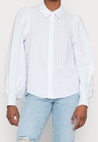 Chemise à rayures bleu clair et blanches, boutonnée, avec des manches bouffantes, un col droit et des poignets ajustés, fabriquée en tissu léger.