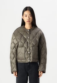 Giacca trapuntata verde oliva con collo rotondo, bottoni a pressione e due tasche frontali. Presenta una texture morbida e lucida e un design imbottito.